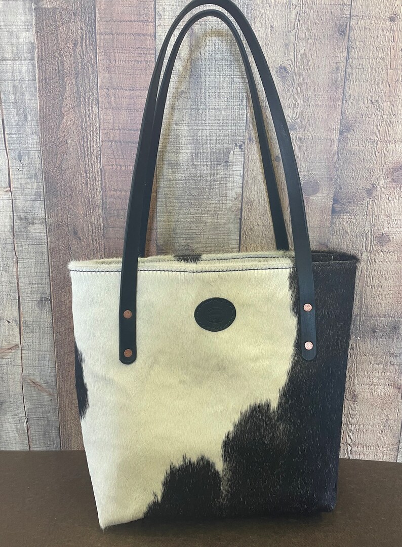 Pu&ograve; includere: Una borsa tote in pelle di mucca bianca e nera con manici in pelle nera e rivetti in rame. La borsa ha una toppa ovale in pelle nera sul davanti.