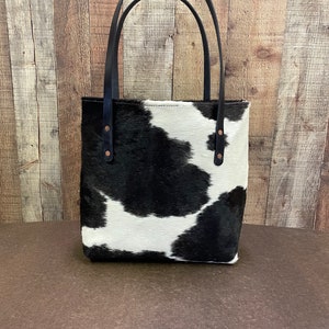 Pu&ograve; includere: Una borsa tote in pelle di mucca bianca e nera con manici in pelle nera e rivetti in rame. La borsa ha una forma rettangolare ed &egrave; realizzata in pelle morbida e flessibile.