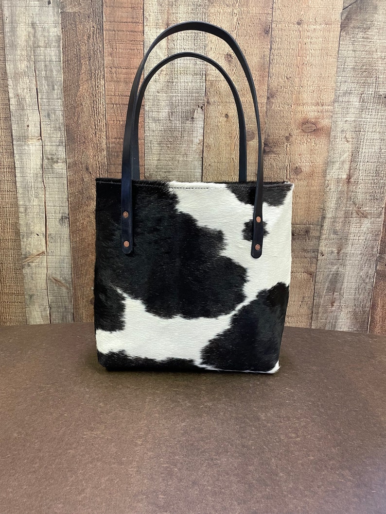 Pu&ograve; includere: Una borsa tote in pelle di mucca bianca e nera con manici in pelle nera e rivetti in rame. La borsa ha una forma rettangolare ed &egrave; realizzata in pelle morbida e flessibile.