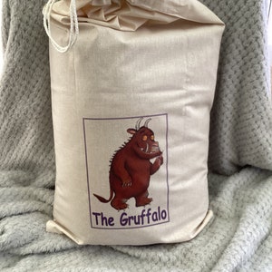 May include: A beige drawstring bag featuring a brown and red illustration of the Gruffalo from the children's book. The bag has the text "The Gruffalo" printed on the front.