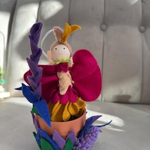May include: A handcrafted felt fairy doll in a terracotta pot, decorated with felt flowers and leaves. The fairy has a yellow crown, a pink dress, and a smiling face. The pot is adorned with blue, green, and purple felt foliage.
