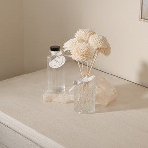 May include: A clear glass bottle with a black lid and a white label with the text "Vanilla" on it. The bottle is sitting on a white stone base. A clear glass vase with a white ribbon and a bouquet of white wooden flowers is next to the bottle.