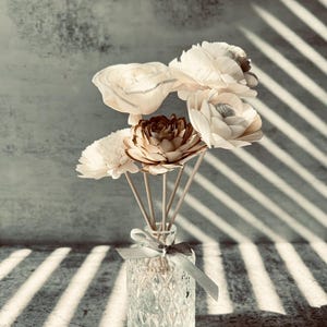 May include: A clear glass diffuser bottle with a textured surface and a gray ribbon. The bottle is filled with wooden sticks and topped with a bouquet of white and brown wooden flowers.