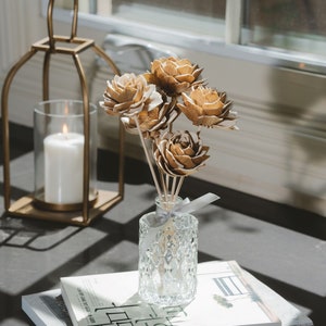 May include: A clear glass diffuser bottle with a textured surface, filled with a light brown liquid and topped with wooden sticks. The bottle is decorated with a light gray ribbon. The diffuser is surrounded by a bouquet of dried, light brown flowers.