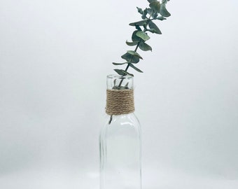 May include: A clear glass vase with a square base and a natural twine tied around the neck. A single sprig of dried eucalyptus is in the vase.