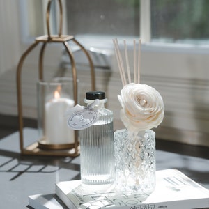 May include: A clear glass diffuser bottle with a black cap and a white flower on top. The diffuser is sitting on a stack of books with Korean text. The diffuser is surrounded by natural light.