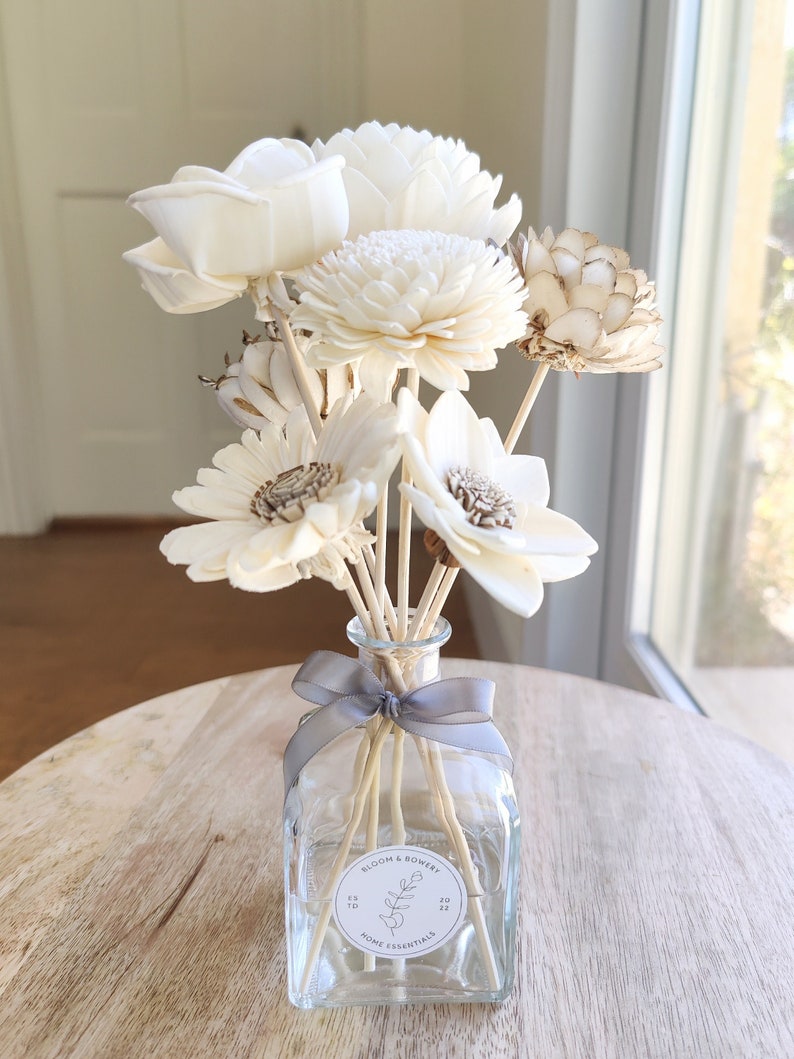May include: A glass diffuser bottle with a grey ribbon and wooden sticks. The bottle has a white label with the text "Bloom & Bowery Home Essentials" and a floral design. The bottle is surrounded by a bouquet of dried white flowers.