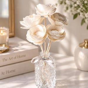May include: A clear glass diffuser bottle with a diamond-patterned texture and a light gray ribbon tied around the neck. Five cream-colored, layered wooden flowers are attached to thin wooden reeds, extending upward from the bottle opening.
