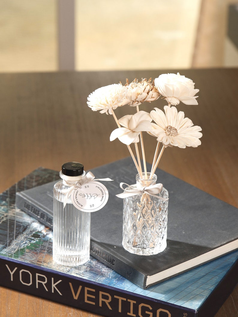 May include: A clear glass bottle with a black lid and a white label that says "Bloom & Botany" and "Stylized Branch" is sitting on a stack of books. The bottle is filled with a clear liquid and has a white ribbon tied around the neck. A clear glass diffuser bottle with a white ribbon is sitting on top of the books. The diffuser bottle is filled with a clear liquid and has a bouquet of dried white flowers in it.