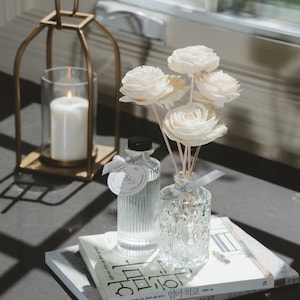 May include: A white diffuser bottle with a clear glass diffuser stick and a bouquet of white wooden flowers. The diffuser bottle is sitting on a stack of books with Korean text.