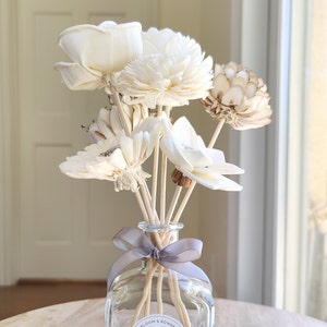 May include: A clear glass diffuser bottle with a grey ribbon bow and wooden sticks. The bottle is filled with dried white flowers and a few brown flowers.