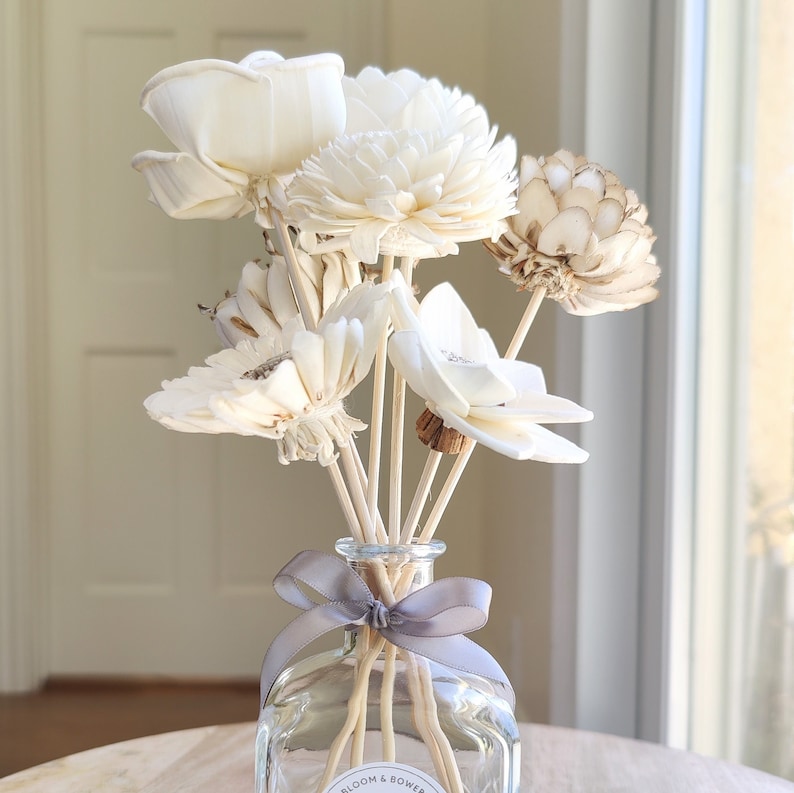 May include: A clear glass diffuser bottle with a grey ribbon bow and wooden sticks. The bottle is filled with dried white flowers and a few brown flowers.