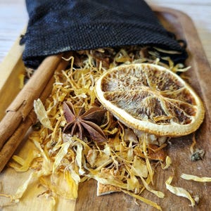 Op de afbeelding: Een zwarte stoffen zak gevuld met gedroogde citrusschillen, steranijs en kaneelstokjes. De ingrediënten zijn verspreid over een houten oppervlak.
