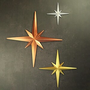 May include: Three metallic starburst wall decor pieces in copper, silver, and gold tones. The stars vary in size and are arranged against a dark gray textured background. The largest star is copper.