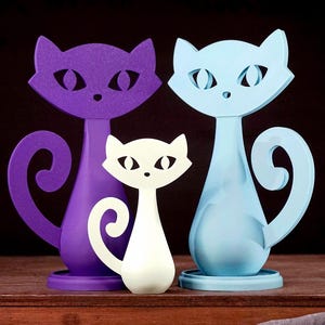 May include: Three stylized cat figurines in purple, white, and light blue. The cats have cut-out eyes and curled tails. The figurines are displayed on a wooden surface against a dark background.