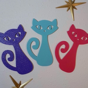 May include: Three colorful cat wall decorations in purple, turquoise, and red. Each cat has a stylized design with cut-out eyes and a curled tail. Gold starburst ornaments are also visible.