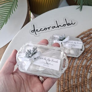 May include: Two small, ornate silver trays, each holding white candies, are presented. The trays are adorned with white tulle, bows, and tags that read "Gizem & Alp Burak." The background includes greenery and a woven mat.