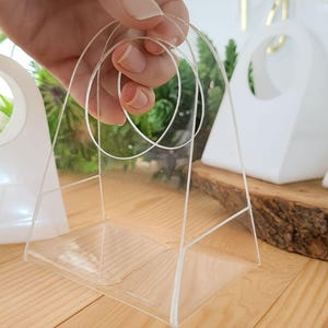 May include: Clear acrylic geometric display box with a handle. The box is held by a hand, showcasing its transparent design. The box is on a wooden surface, with other white geometric shapes in the background.