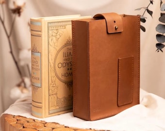 May include: A tan leather book cover and a vintage book titled "The Iliad and The Odyssey of Homer" are displayed on a wooden surface. The book cover has a flap closure with a brass button. The book has a cream-coloured cover with gold lettering.