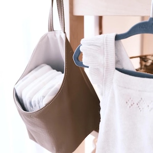 May include: A taupe hanging storage pouch with a white interior lining, filled with a stack of folded white diapers. The pouch features a single top handle and is suspended from a hook. Beside it hangs a white sleeveless garment on a blue hanger.