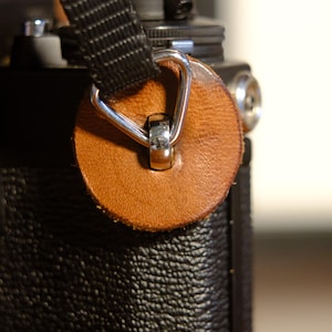 Op de afbeelding: Close-up van een camera met een zwarte riem en een bruine leren bevestiging. Het leren stuk is cirkelvormig met een zilveren metalen ring. De camerabehuizing is zwart met een textuur.