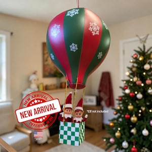 May include: A hanging hot air balloon decoration featuring alternating red and green vertical panels with white snowflake patterns. A square basket with a green and white checkerboard pattern hangs below, containing two small elf dolls.