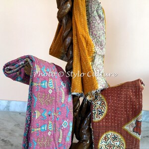 May include: Three kantha quilts, a traditional Indian hand-stitched textile, are displayed on a wooden stand. The quilts are made of recycled saris and feature colorful floral and geometric patterns. The quilts are in shades of pink, orange, and brown.