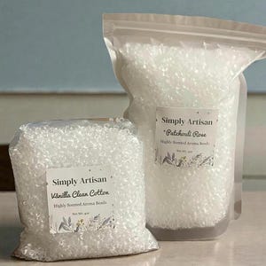 May include: Two clear plastic bags filled with white aroma beads. The bags have labels that read "Simply Artisan" with scent names "Vanilla Clean Cotton" and "Patchouli Rose". The labels also state "Highly Scented Aroma Beads".