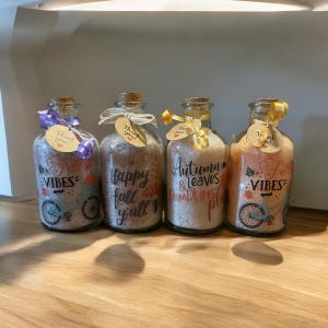 May include: Four glass bottles filled with bath salts, each with a cork stopper and a decorative tag. The bottles have labels with the words "Vibes", "Happy fall y'all", "Autumn & Leaves Pumpkins", and "Fall Vibes". Each bottle is decorated with a ribbon.