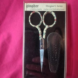 May include: Gingher Designer's Series Lindsay 4-inch embroidery scissors in a clear plastic case. The scissors have a floral pattern on the handles and come with a black leather sheath. The packaging includes the text "a tradition of quality."
