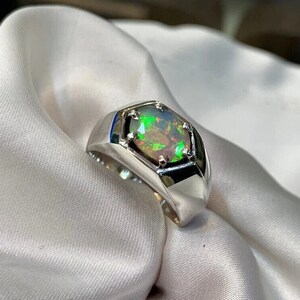 Pode incluir: Um anel de prata com uma configuração hexagonal apresentando uma gema de opala verde, preta e iridescente.