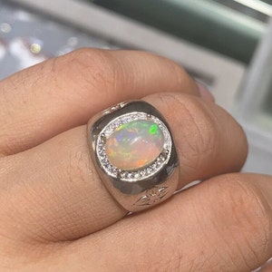 Może przedstawiać: Srebrny pierścionek z dużym owalnym opalem otoczonym mniejszymi, przezroczystymi kamieniami szlachetnymi. Pierścionek ma ozdobny wzór kwiatowy na obrączce.