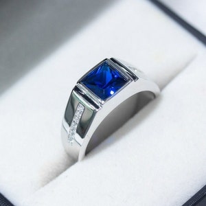 Op de afbeelding: Een zilveren ring met een vierkante blauwe edelsteen en kleine heldere edelstenen op de band.