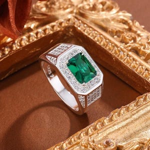 May include: A silver ring with a rectangular, emerald-cut green gemstone at the center, surrounded by smaller, clear stones. The ring has a detailed design on the band and around the central stone, with a polished finish.