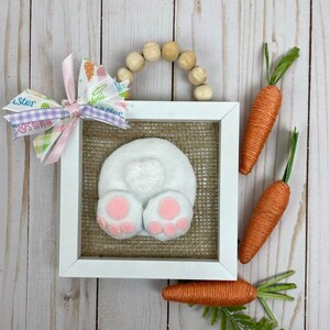 May include: A white bunny butt with pink feet in a white frame with a burlap background. The frame is decorated with a wooden bead garland and a pink and purple ribbon with the words "Easter Blessings". Three orange yarn carrots with green stems are on a white wood background.
