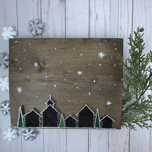 May include: A wooden sign with a string art design of a winter village silhouette. The sign is painted brown and features white stars and snowflakes. The village silhouette is made of black and green string.