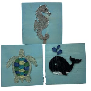 May include: Three square wooden plaques with sea creature designs. The top plaque features a seahorse, the bottom left a turtle, and the bottom right a whale. Each design is made with silver-colored string on a light blue background.