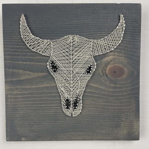 May include: A handcrafted string art piece featuring a bull skull design. The skull is made of silver-colored string, with black accents for the eyes and nose, mounted on a dark gray wooden board. A rustic, western-themed wall decor.