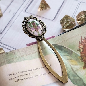 Könnte beinhalten: Antiker Messing-Lesezeichen mit ovaler Kamee mit Feen-Design. Die Fee hat grüne Flügel und ein rosa Kleid. Das Lesezeichen steckt in einem Buch mit dem Text: "Petal gowns and acorn caps so last summer!"