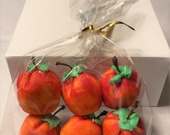 Puede incluir: Una bolsa transparente de celofán atada con una cinta dorada contiene seis melocotones de mazapán. Los melocotones son de color naranja y rojo con hojas verdes y tallos marrones. Una caja blanca está en el fondo.