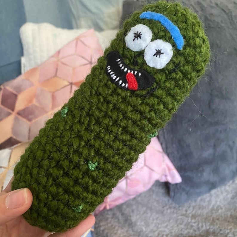 May include: Green crocheted pickle plush toy with embroidered eyes, a mouth, and a tongue. The pickle has a blue felt band around it.