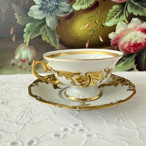 May include: White teacup and saucer set with gold trim and ornate gold detailing. The teacup has a curved handle and sits on a matching saucer. The set is likely porcelain and is a vintage or antique item.