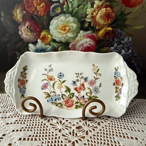 May include: A white rectangular serving platter with a floral design featuring red, blue, and yellow flowers. The platter has a scalloped edge and is supported by a bronze metal stand. The background features a painting of colorful flowers.