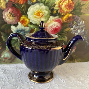 May include: A cobalt blue ceramic teapot with gold trim and accents. The teapot has a ribbed design and a matching lid. The background features a floral painting with various colorful flowers. The teapot is sitting on a white lace tablecloth.