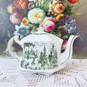 May include: A white ceramic teapot featuring a green transferware pattern depicting a coastal scene with sailing ships, castles, and trees. The teapot has an octagonal shape, a matching lid with a small finial, and green trim on the handle and spout.