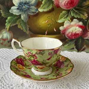 May include: A vintage teacup and saucer set. The cup and saucer are decorated with a green background and a floral pattern of pink and yellow roses. The rims are trimmed with gold. The teacup has a white interior and a curved handle.