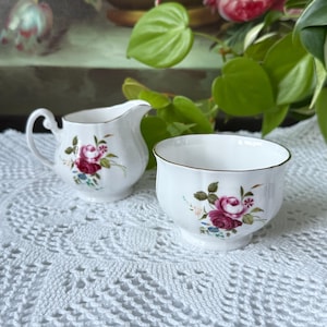 May include: A white porcelain creamer and sugar bowl set. Each piece is decorated with a floral pattern featuring pink roses, green leaves, and gold trim. The creamer has a handle and spout, while the bowl has a scalloped edge.