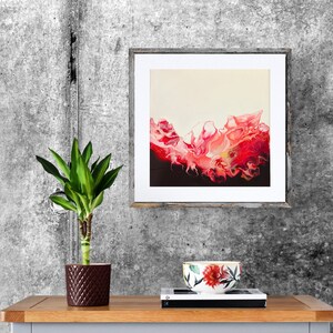 May include: A framed abstract painting with a pink and red wave-like design on a cream background. The painting is hanging on a grey concrete wall above a wooden table with a potted plant and a floral patterned cup.