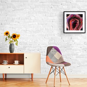 May include: A white brick wall with a framed abstract painting of pink, red, and black swirls. A wooden chair with a patchwork fabric seat and a white and brown cabinet with two cups on the top shelf are in the foreground.