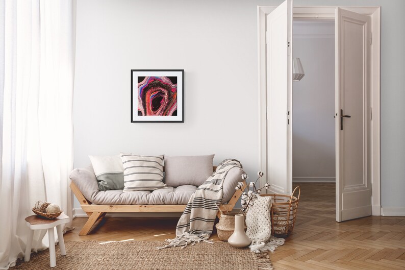 May include: A framed abstract painting with pink, red, blue, and black colors hangs on a white wall above a light gray couch with a striped throw blanket and pillows. The couch is in a living room with a wooden floor and a white door.